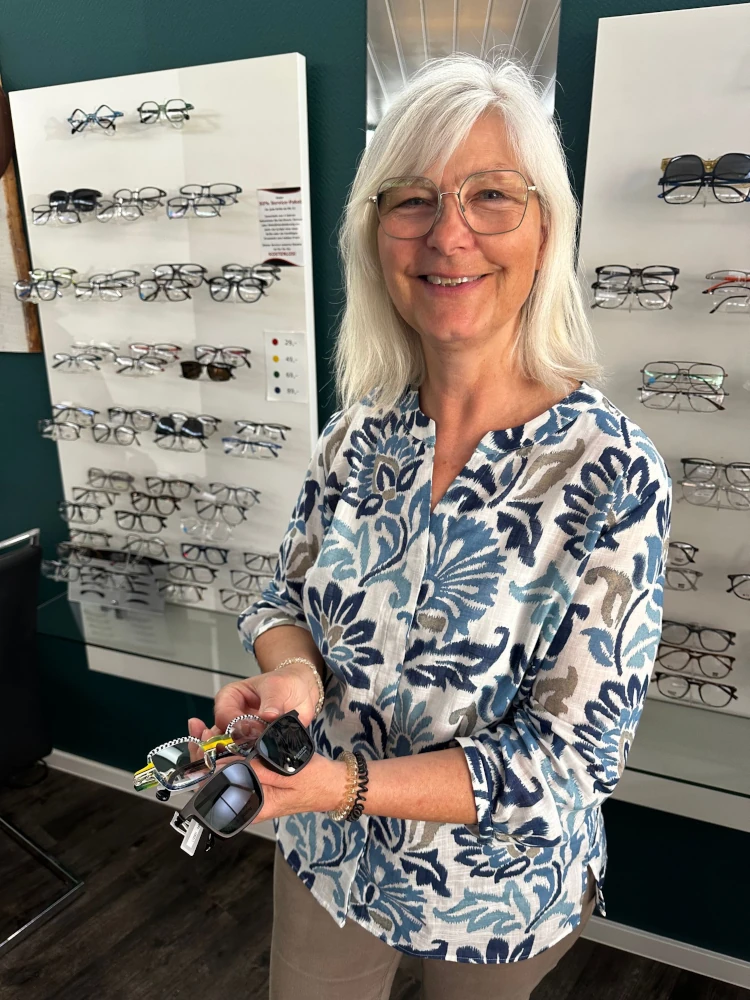 Ältere Frau mit Brille steht in einem Optikerladen vor einer Auswahl an Brillengestellen.