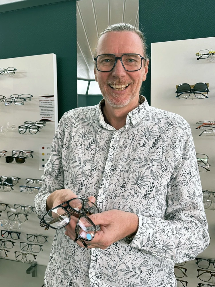 Ein Mann mit Brille steht lächelnd im Optikergeschäft und hält mehrere Brillengestelle.