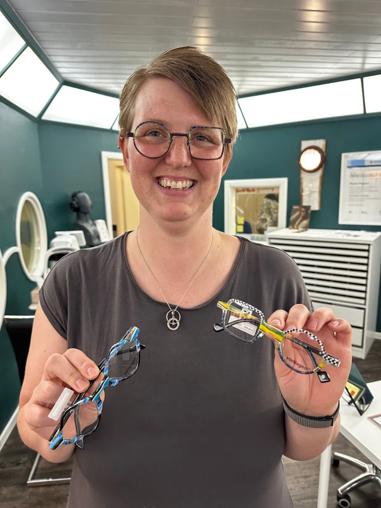 Lächelnde Frau im Brillengeschäft hält farbenfrohe Brillengestelle zur Auswahl in den Händen.